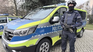 Umundurowany i uzbrojony policjant Grupy szybkiego reagowania przy oznakowanym radiowozie Renault Traffic