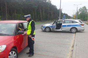 Policjant dokonujący pomiaru stanu trzeźwości kierowcy jadącego czerwoną osobówką. W tle policyjny radiowóz.