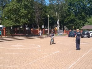 dziecko jedzie rowerem po wyznaczonych kredą liniach na boisku szkolnym. W tle policjantka