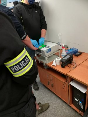 Policjant wydziału kryminalnego Komisariatu Policji Toruń Śródmieście pilnujący podejrzanego o posiadanie znacznej ilości narkotyków podczas pobierania śladów  daktyloskopijnych z użyciem urządzenia skanującego.