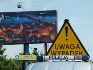 Zdjęcie poglądowe do wypadku - galeryjka z napisem policja na policyjnym radiowozie oraz znak uwaga wypadek, na tle plakatu Toruń.