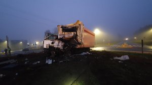 Zdjęcie z miejsca wypadku dwóch ciężarówek, jedna z nich z pomarańczową naczepą zniszczona stoi  na środku na wysepce z zielenią w środku ronda czyli skrzyżowania z ruchem okrężnym.