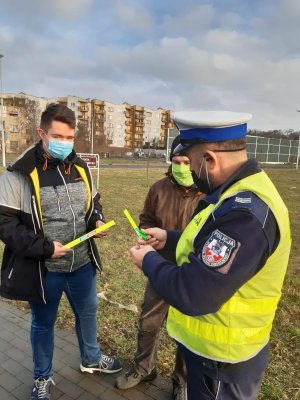 policjant stoi tyłem. frontem do niego dwójka mężczyzn. Wszyscy trzymają opaski odblaskowe