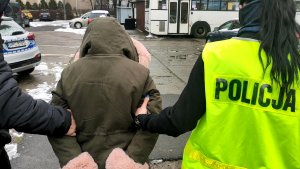 Policjantka w kamizelce odblaskowej z napisem policja wraz z policjantem służb kryminalnych, który na lewym ramieniu ma opaskę z napisem policja prowadzi zatrzymaną, oszustkę, która udawała policjantkę. Zatrzymana ubrana jest w kurtkę w kolorze khaki, czarne spodnie i czarne kozaki. Na głowie ma założony kaptur obszyty jasnoróżowym futerkiem . Zatrzymana dłoniach, które są z tyłu ma założone kajdanki. Funkcjonariusze przechodzą parkingiem znajdującym się na podwórku Komisariatu Policji Toruń Śródmieście.