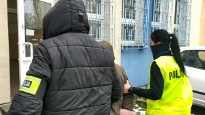 Policjantka w kamizelce odblaskowej z napisem policja wraz z policjantem służb kryminalnych, który na lewym ramieniu ma opaskę z napisem policja prowadzi zatrzymaną, oszustkę, która udawała policjantkę. Zatrzymana ubrana jest w kurtkę w kolorze khaki, czarne spodnie i czarne kozaki. Na głowie ma założony kaptur obszyty jasnoróżowym futerkiem . Zatrzymana dłoniach, które są z tyłu ma założone kajdanki. Funkcjonariusze przechodzą parkingiem znajdującym się na podwórku Komisariatu Policji Toruń Śródmieście. Wszyscy wchodzą przez w połowie oszklone drzwi do budynku  jednostki.