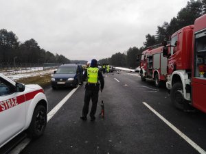 droga, na poboczu po prawej i lewej stronie wozy strażackie, w środku kadru policjant, w tle rozbite samochody