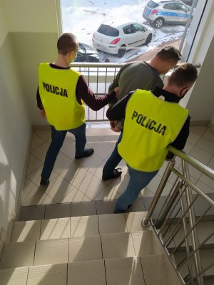 Dwaj policjanci w kamizelkach służbowych z napisem Policja  prowadzą  na przesłuchanie zatrzymanego klatką schodową  w Komisariacie Policji Toruń Podgórz.