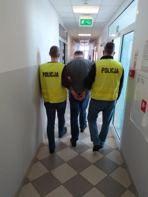 Dwaj policjanci w kamizelkach służbowych z napisem Policja  prowadzą  na przesłuchanie zatrzymanego korytarzem  w Komisariacie Policji Toruń Podgórz.