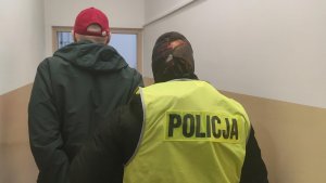 Policjant kryminalny ubrany w kamizelkę służbową z napisem Policja prowadzi zatrzymanego korytarzem  w Komisariacie Policji Toruń Śródmieście. Zatrzymany 28 latek ubrany jest  w szarą bluzę, ciemne spodnie a na głowie ma czerwoną czapeczkę bejsbolową. Na dłoniach na których ma fioletowe lateksowe rękawiczki ma założone kajdanki, które są z tyłu.