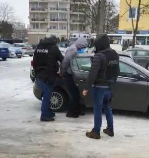 Dwaj kryminalni z Komisariatu Policji Toruń Śródmieście ubrani w czarne kamizelki z napisem policja wychodzą z budynku jednostki na wewnętrzny parking wraz za zatrzymanym, który ubrany jest w szarą kurtkę z kapturem i założonymi na dłoniach z tyłu kajdankami. Podejrzany ma na głowie założony kaptur. Podejrzany wsiada do radiowozu. Jeden z policjantów trzyma podejrzanego a drugi otwiera mu drzwi do służbowego, nieoznakowanego  radiowozu.