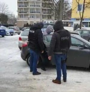 Dwaj kryminalni z Komisariatu Policji Toruń Śródmieście ubrani w czarne kamizelki z napisem policja wychodzą z budynku jednostki na wewnętrzny parking wraz za zatrzymanym, który ubrany jest w szarą kurtkę z kapturem i założonymi na dłoniach z tyłu kajdankami. Podejrzany ma na głowie założony kaptur. Podejrzany wsiada do radiowozu.