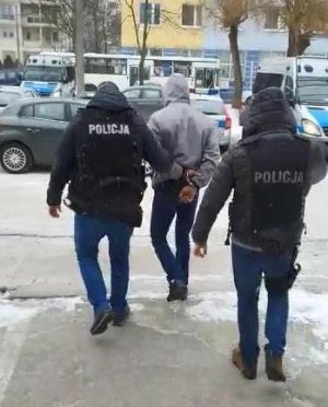 Dwaj kryminalni z Komisariatu Policji Toruń Śródmieście ubrani w czarne kamizelki z napisem policja wychodzą z budynku jednostki na wewnętrzny parking wraz za zatrzymanym, który ubrany jest w szarą kurtkę z kapturem i założonymi na dłoniach z tyłu kajdankami. Podejrzany ma na głowie założony kaptur.