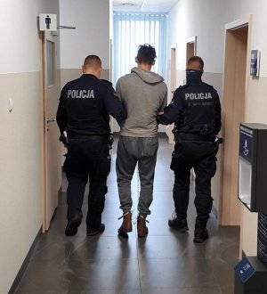 Policjanci prewencji ubrani w granatowe mundury prowadzą sprawcę zniszczenia radiowozów korytarzem Komisariatu Policji w Dobrzejewicach do pokoju przesłuchań. Podejrzany ma na dłoniach i stopach kajdanki.