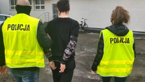 Policjanci kryminalni w kamizelkach służbowych prowadzą podejrzanego, który ma założone kajdanki na dłonie z tyłu