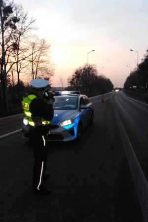Zdjęcia wieczorem i nocą policjanta podczas działań na drodze krajowej numer 91. Policjant w kamizelce i z ręcznym miernikiem prędkości.