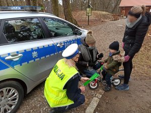 policjantka w kamizelce odblaskowej kuca przy radiowozie i podaje dziecku na rowerku biegowym opaskę odblaskową. Przy dziecku stoją dwie kobiety