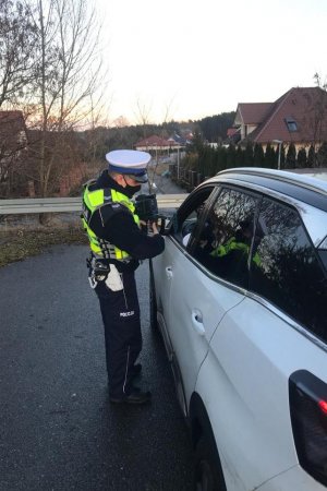 Zdjęcia funkcjonariusza Wydziału Ruchu Drogowego na drodze z ręcznym miernikiem prędkości  oraz zatrzymany pojazd . Policjant pochylający się przy pojeździe koloru białego i pokazujący kierowcy zarejestrowany jego pomiar prędkości na mierniku.