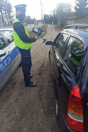 Policjant podczas kaskadowego pomiaru prędkości  trzymający urządzenie w dłoni i stojący przy drodze. Odczyt z monitora videorejestratora.