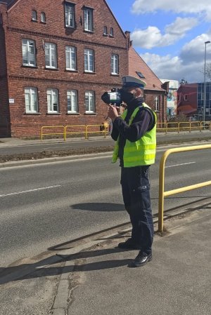 Policjant podczas kaskadowego pomiaru prędkości  trzymający urządzenie w dłoni i stojący przy drodze. Odczyt z monitora videorejestratora.