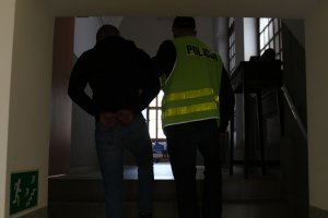 Zdjęcie oszusta internetowego  prowadzonego  korytarzem w  Komisariacie Policji Chełmży przez policjanta na przesłuchanie. Podejrzany ubrany w czarną bluzę i spodnie dżinsowe. Policjant w neonowej, odblaskowej kamizelce z napisem POLICJA. Obaj w maseczkach.