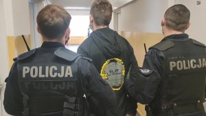 Zdjęcie podejrzanego prowadzonego korytarzem komisariatu  przez dwóch umundurowanych policjantów prewencji.