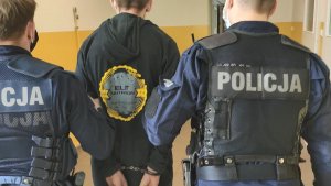 Zdjęcie podejrzanego prowadzonego korytarzem komisariatu  przez dwóch umundurowanych policjantów prewencji.