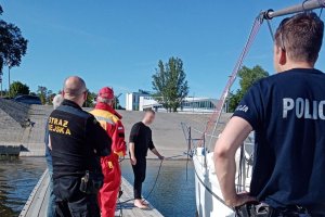 przystań dla łodzi, na pomoście umundurowany policjant, strażnik miejski, ratownik WOPR oraz mężczyzna trzymający linę dołączoną do łodzi, łódź stoi przy pomoście