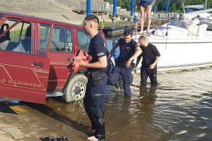 przystań dla łodzi, na pochylni prowadzącej do nurtu stoi samochód, do którego przyczepiona jest żaglówka, dwójka policjantów oraz mężczyzna ubrany na czarno brodzą w płytkiej wodzie