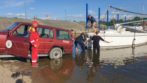 przystań dla łodzi, na pochylni prowadzącej do nurtu stoi samochód, do którego przyczepiona jest żaglówka, dwójka policjantów oraz mężczyzna ubrany na czarno brodzą w płytkiej wodzie, na lądzie stoi ratownik WOPR