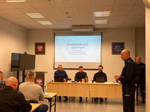 Widoczny stojący policjant w umundurowaniu oraz stojący strażak w umundurowaniu, a także mężczyźni siedzący w ubraniach cywilnych. W tle tablica z napisem poszukiwania osób zaginionych Wydział kryminalny Komendy Miejskiej Policji w  Toruniu Zespół Poszukiwań i Identyfikacji Osób