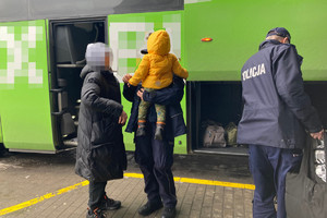 umundurowana policjantka trzyma na rękach dziecko, obok stoi chłopiec w kapturze na głowie, z boku stoi policjant, w tle widoczny autokar