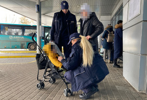 umundurowana policjantka kuca przy wózku w którym jest małe dziecko, obok stoi chłopiec w kapturze na głowie i umundurowany policjant