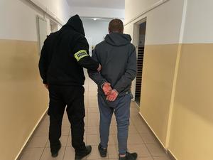 osoba w bluzie z kapturem na głowie i opaska policja na ramieniu trzyma zakutego w kajdanki mężczyznę