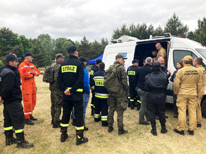 strażacy, żołnierze i nieumundurowani stoją w kolejce do pojazdu dowodzenia z napisem policja