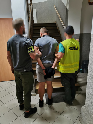 dwóch nieumundurowanych policjantów prowadzi mężczyznę w kajdanckach