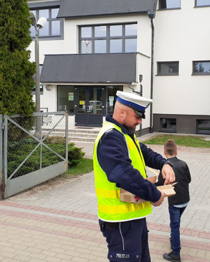 policjant przed budynkiem szkoły wręcza dziecku opaskę odblaskową