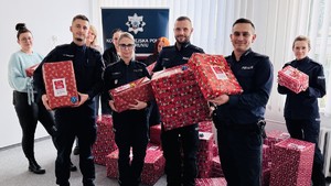 Na zdjęciu widać pracowników cywilnych oraz policjantów. Trzymają w rękach prezenty. W tle znajduje się rolap Komendy Miejskiej Policji w Toruniu.