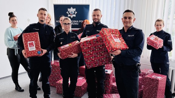 Na zdjęciu widać pracowników cywilnych oraz policjantów. Trzymają w rękach prezenty. W tle znajduje się rolap Komendy Miejskiej Policji w Toruniu.