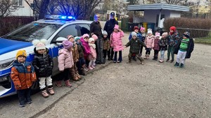 Na zdjęciu widzimy przedszkolaków z opiekunem oraz policjantką. Stoją na tle policyjnego radiowozu.