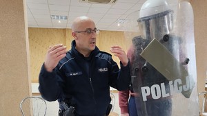 Policjant opowiada o swojej pracy. W tle widać ubranego w sprzęt PZ uczestnika spotkania.