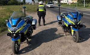 Na zdjęciu widać umundurowanego policjanta ruchu drogowego stojącego przy policyjnym motocyklu. Zdjęcie wykonano w rejonie przejazdu kolejowego.