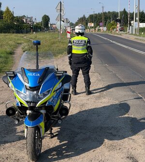 Na zdjęciu widać umundurowanego policjanta ruchu drogowego stojącego przy policyjnym motocyklu. Zdjęcie wykonano w rejonie przejazdu kolejowego.