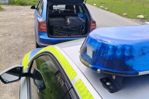 Na pierwszym zdjęciu znajduje się oznakowany radiowóz policyjny oraz bagażnik pojazdu zatrzymanego do kontroli, w którym w przestrzeni bagażowej znajdują się  pudła owinięte czarną folią. Na kolejnym zdjęciu widać nieumundurowanego policjanta, znajdującego się przy zabezpieczonych nielegalnych papierosach.