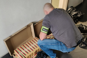 Na pierwszym zdjęciu znajduje się oznakowany radiowóz policyjny oraz bagażnik pojazdu zatrzymanego do kontroli, w którym w przestrzeni bagażowej znajdują się  pudła owinięte czarną folią. Na kolejnym zdjęciu widać nieumundurowanego policjanta, znajdującego się przy zabezpieczonych nielegalnych papierosach.